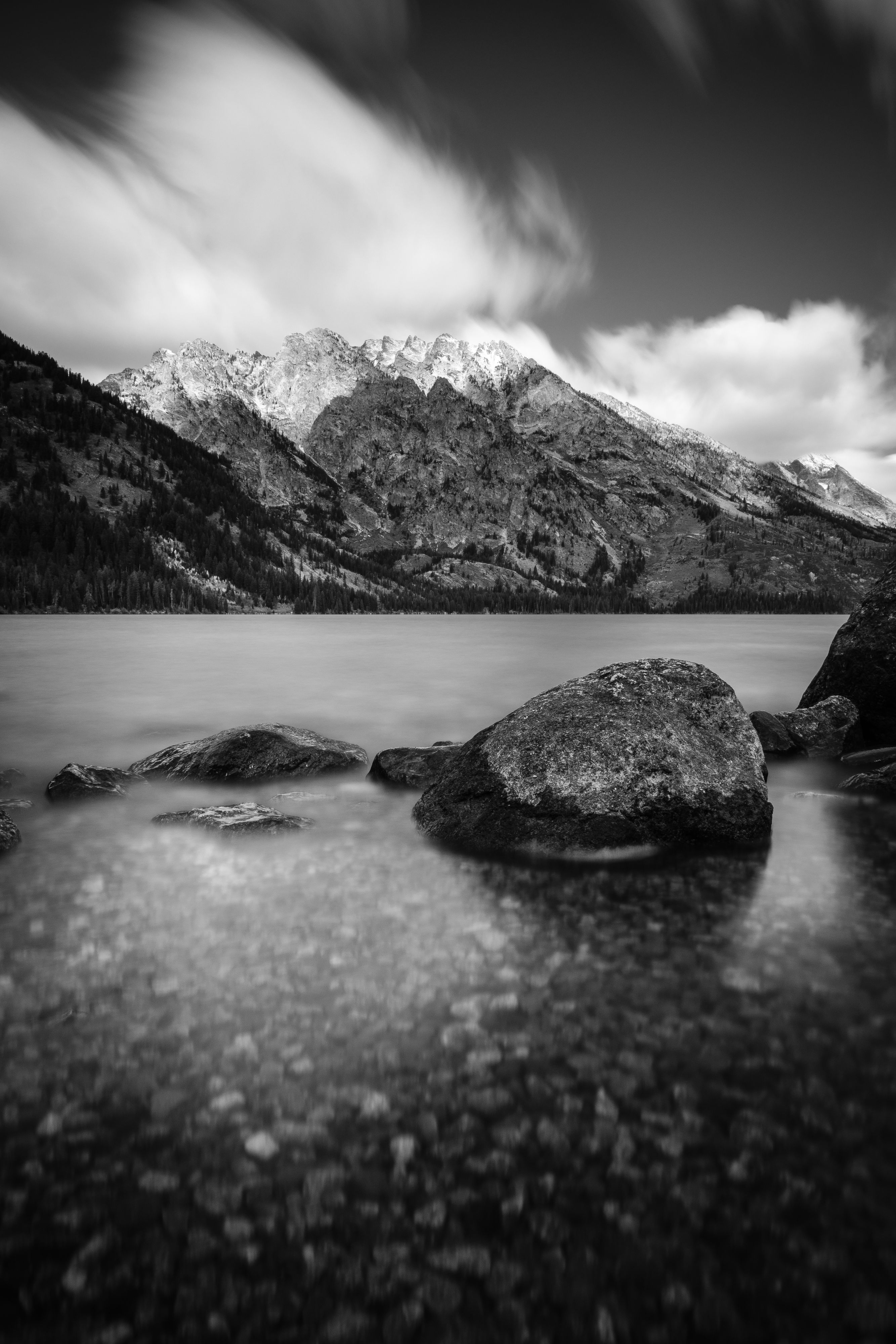 A long exposure of the shores of Jenny Lake in Grand Teton National Park. A number of large boulders can be seen in the blurry water in the foreground. In the background, Mount Saint John, partially lit by the sun, and clouds in the sky blurred by their motion.