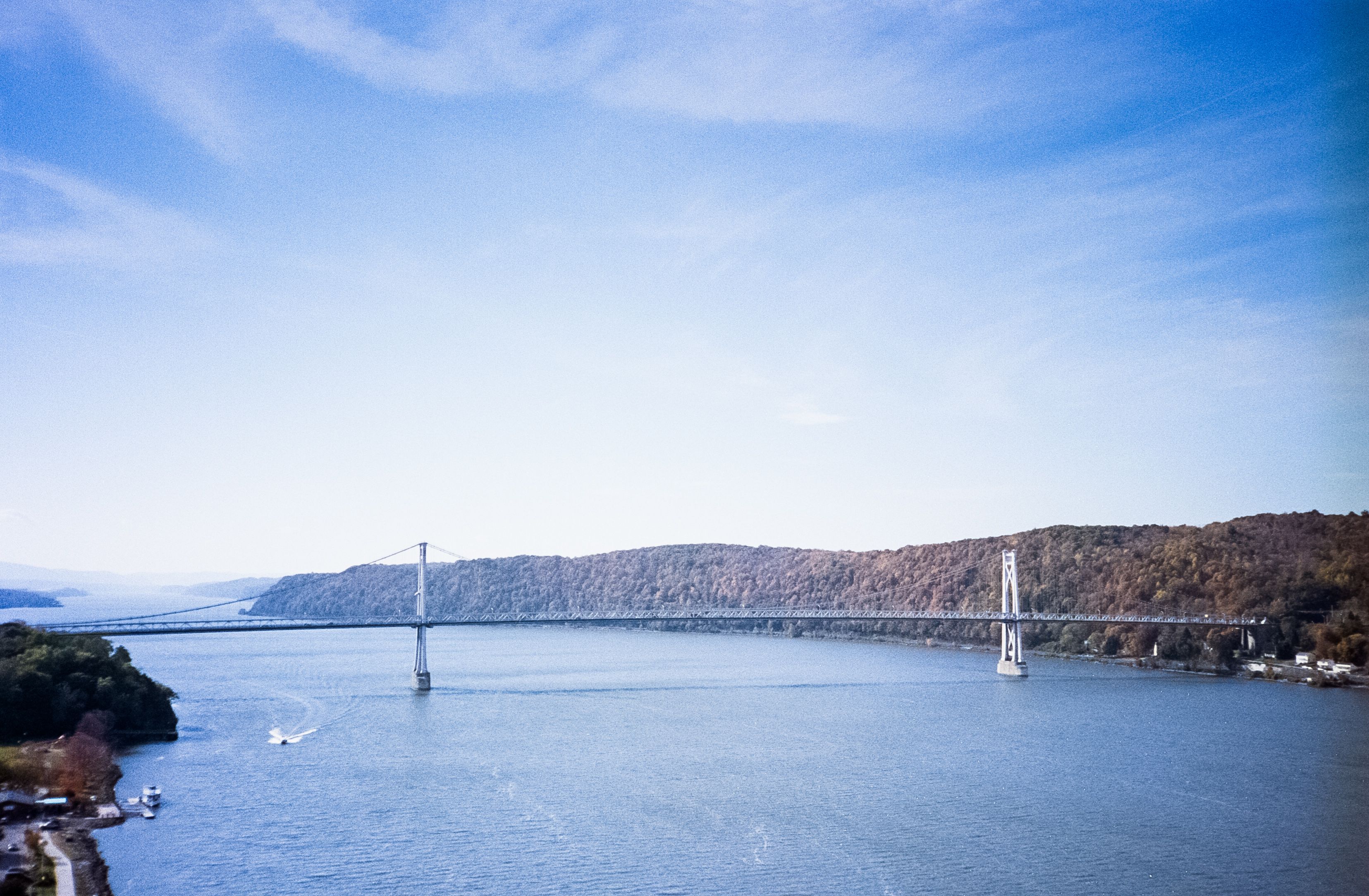 The Franklin Delano Roosevelt Mid-Hudson Bridge, a long suspension bridge spanning across the Hudson River, connecting two forested shores. The landscape includes hills covered with trees, and a few boats are visible on the river. The sky is clear with some clouds.